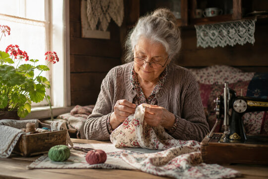 Elderly woman sewing floral fabric at sunlit window in cozy craft room, using vintage sewing machine on wooden table with yarn balls and wicker basket