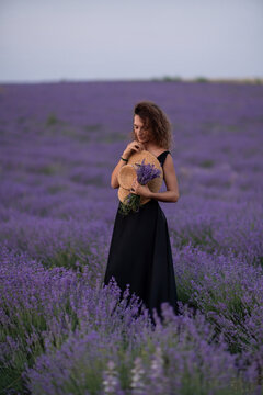 Elegant woman in black dress holds straw hat and Lavandula angustifolia bouquet within vast blooming lavender field at dusk