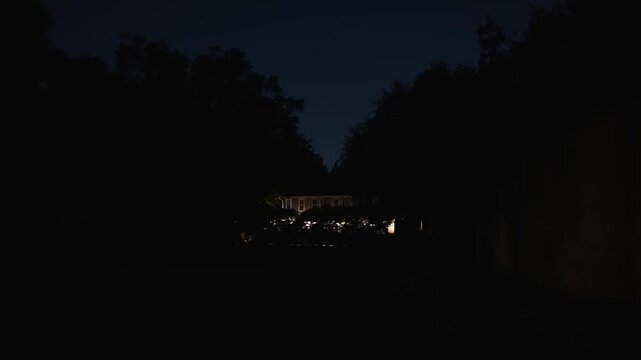 Lonely residential house barely visible among dark trees at night, with faint light revealing the building deep within an almost completely dark neighborhood