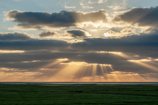 Sunbeams pierce dark clouds over a green coastal landscape at sunset
