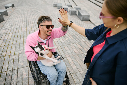 Man in wheelchair holding dog gives high five to woman in city