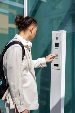 Side view of a traveler with backpack pressing buttons on an entry intercom panel beside a glass door of a residential building. Concept of apartment access control and self check-in arrival.