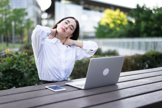 Businesswoman stretches her neck while working on laptop outdoors