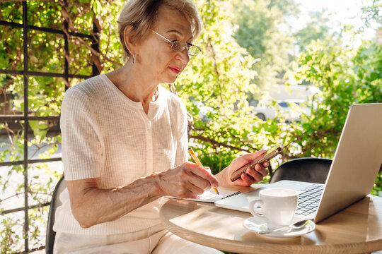 Woman writes in notebook with smartphone and laptop at outdoor cafe