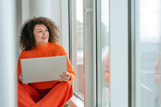 Smiling woman with laptop in orange sweater sitting on windowsill