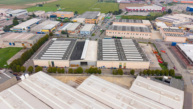 Aerial view of an industrial complex featuring large warehouses with flat roofs and skylights, surrounded by paved areas and green fields in Milan, Lombardy, Italy.