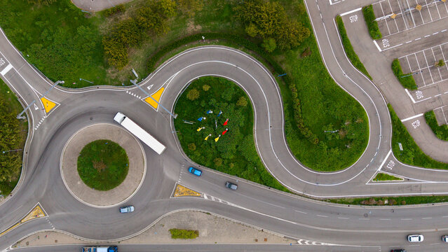 Aerial view of a roundabout in a commercial zone with a white truck and cars navigating the intersection surrounded by green landscaping in San Giuliano Milanese, Lombardy, Italy.