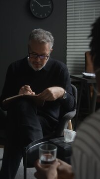 Vertical shot of middle aged Caucasian therapist listening to patient and writing notes during session. Footage suitable for mental health campaigns, psychotherapy apps, and counseling advertising