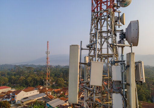 Aerial view of a telecommunications tower equipped with various antennas and dishes overlooking a residential area with lush trees in Cilongok, Central Java, Indonesia.