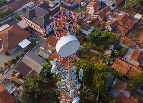 Aerial view of a telecommunications tower with microwave dishes and antennas rising above a residential neighborhood with terracotta rooftops and lush trees in Cilongok, Central Java, Indonesia.