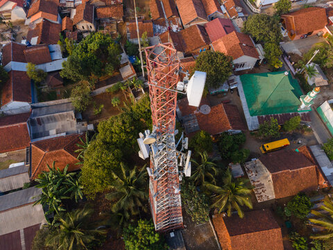 Aerial view of a telecommunications tower surrounded by residential houses with terracotta roofs, lush palm trees, and a mosque in Cilongok, Central Java, Indonesia.