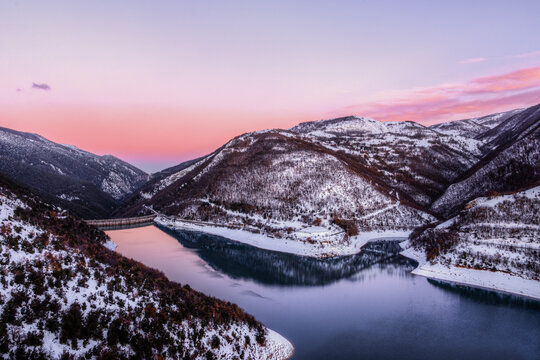 View of the snow-covered mountains surrounding the calm waters of Lake Fiastra during a vibrant sunset in Fiastra, Marche, Italy.