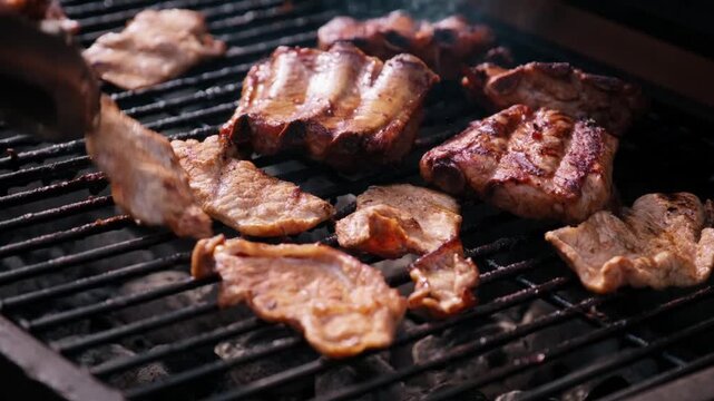 Close up of tongs flipping grilled pork steaks and ribs cooking on a hot barbecue grate during a backyard cookout.