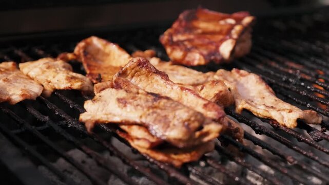 Close up of tongs flipping grilled pork steaks and ribs cooking on a hot barbecue grate during a backyard cookout.