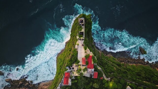 Uluwatu temple and surrounding buildings are built on a dramatic cliff, overlooking the powerful breaking waves of the Indian Ocean, a popular tourist landmark in Bali, Indonesia. Top down aerial view