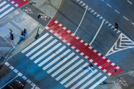 Aerial view of a pedestrian crossing at night with red and white markings, traffic lights, and blurred figures of people walking in Warsaw, Masovian Voivodeship, Poland.