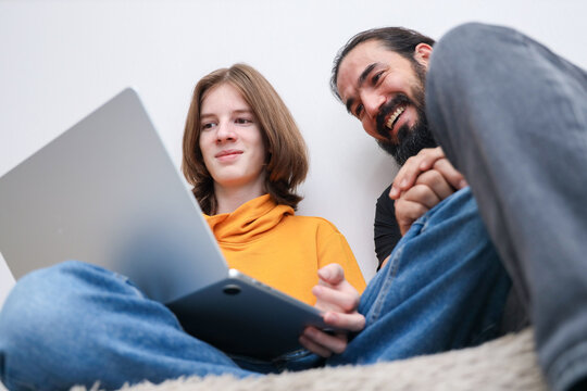 Young Person In Orange Hoodie Sits Beside Bearded Man While Both Look At Tablet Indoors, Showing Relaxed Shared Screen Time, Friendly Guidance, Casual Home Setting