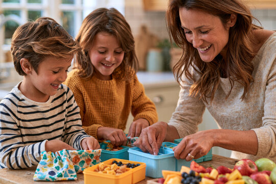 Stylish concept showing smiling parent preparing children's lunches using reusable food containers, cloth snack bags and fresh fruit in a bright family kitchen, warm morning light