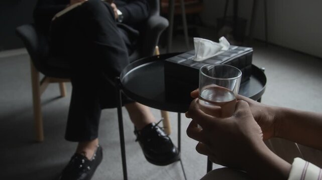 African American young adult woman holding glass of water during psychotherapy session while therapist writes notes. Ideal for mental health campaigns and professional counseling services