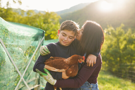 Mother and teenager son having tender moment at organic house garden while holding chicken - Ecologic farm, family people and bonding concept - UGC content