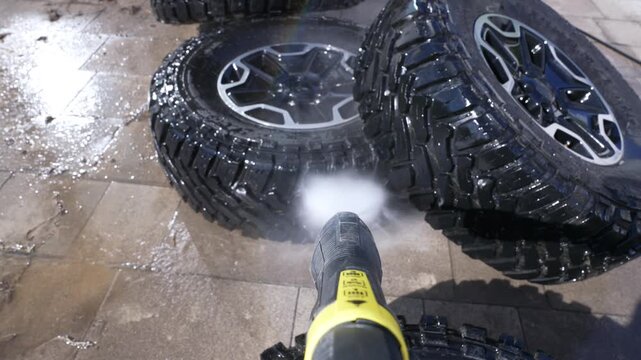 High-pressure water cleans muddy off-road tires. Spray blasts dirt from rugged vehicle wheels. Water droplets splash across wet stone pavement. Scene shows focused, practical cleaning action