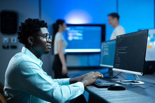 African male software engineer writing program code on multiple desktop screens in a modern office.