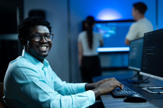 Portrait of African male software engineer writing program code on multiple desktop screens in a modern office.