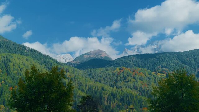 Mountain peaks pine forest autumn landscape with high rocky summits partially covered by white clouds under a blue sky. Scenic alpine wilderness scenery with colorful foliage patches on hillsides, han