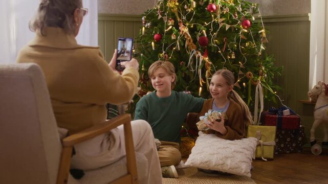 Senior Caucasian woman taking smartphone photo of happy grandchildren holding gift near Christmas tree. Footage suited for seasonal advertising and holiday campaigns
