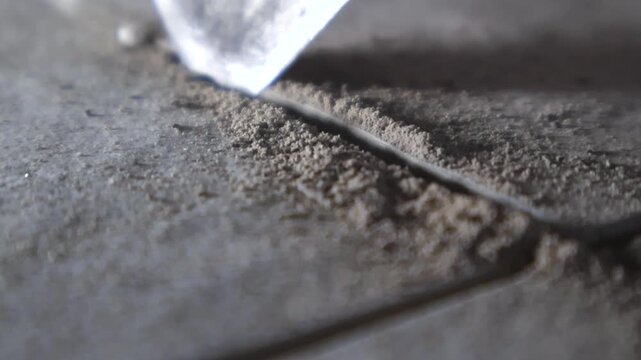 Metal putty knife meticulously scraping and cleaning the hardened old grout from the joint between two dirty ceramic floor tiles during a home renovation or repair project in slow motion