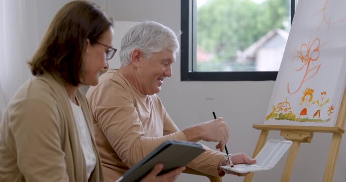 Senior man paints lower canvas details, dips brush into palette, turns toward senior woman during encouragement, then resumes artwork, expressing home art therapy, emotional care, mindful retirement.