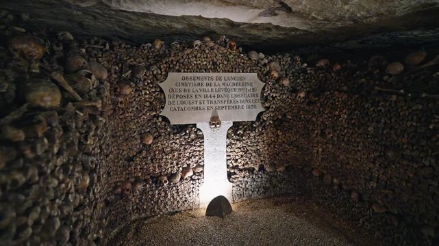 Camera holds on a lit stone marker amid densely stacked bones in a small chamber of the Catacombs of Paris.