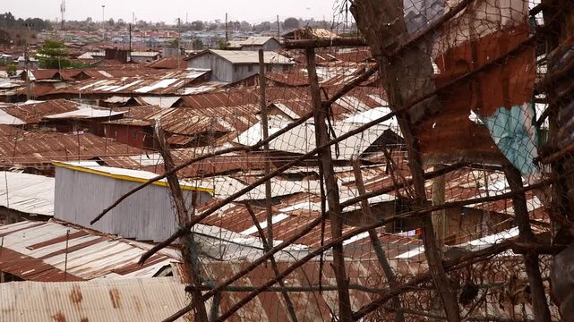 A high-angle view over the dense rusty tin rooftops of Kibera slum in Nairobi, Kenya, seen through a makeshift fence of branches, wire, and fabric.