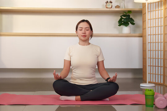 Asian woman meditating in lotus pose on mat at home for mental wellness and yoga fitness