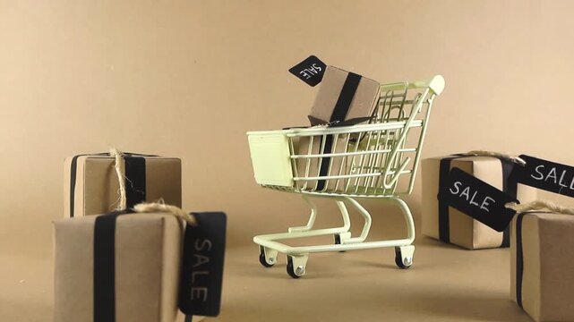 Miniature shopping cart filling up with kraft paper gift boxes with black sale tags against a light brown background, representing a shopping spree during a promotion or holiday season sale