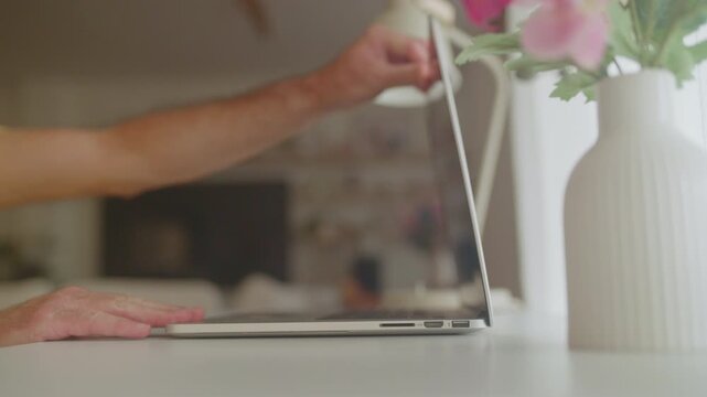 Male hands opening a sleek modern laptop and beginning to type on the keyboard, working from a bright and cozy home office setup with a vase of flowers and a desk lamp nearby