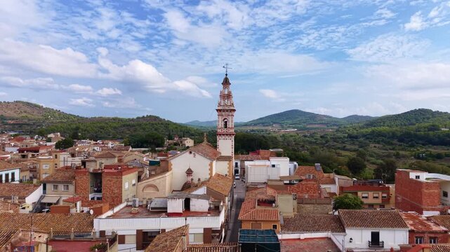 Elevated view of the Church of the Holy Johns in Estivella, Valencia, Spain, with its ornate Baroque bell tower featuring polychrome decoration rising above terracotta rooftops and green hills