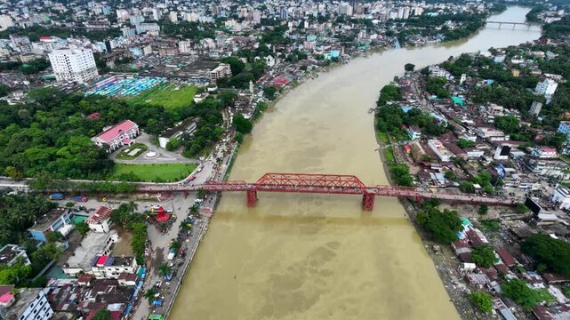 Aerial tracking shot over red steel bridge crossing wide muddy river through dense city with rooftops and green trees, Keane Bridge, Surma River, Sylhet.