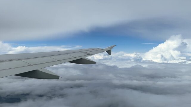 View from airplane window showing aircraft wing above a thick cloud layer with blue sky and cumulus clouds in the background during commercial flight