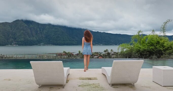 Woman with curly hair wearing blue dress standing near infinity pool edge overlooking Lake Batur and volcanic mountains on a cloudy day in Bali, Indonesia. Travel, vacation, relax, freedom. Back view