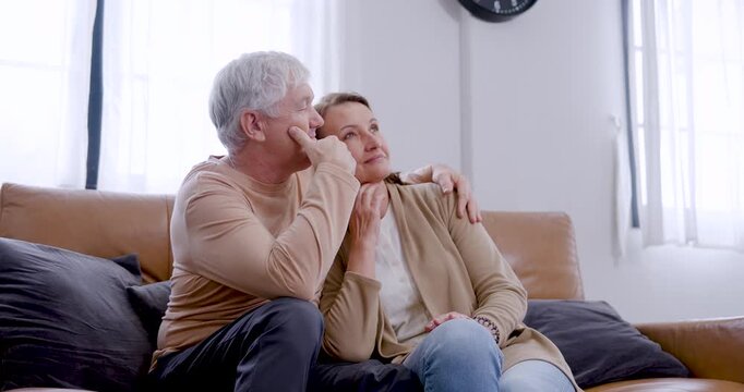 Affectionate senior Caucasian couple cuddle on sofa, rest heads together, gesture while looking upward, and smile softly during peaceful conversation and meaningful connection in later life.