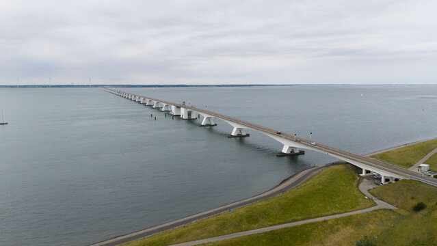 Aerial view of Zeeland Bridge spanning the Oosterschelde estuary with calm waters, green coastal embankments, and a cloudy sky in Zierikzee, Zeeland, Netherlands.