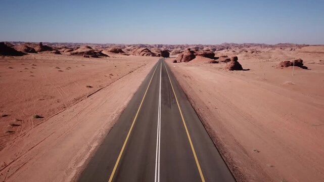 a long asphalt highway stretches straight through a vast, arid desert landscape dotted with rugged rock formations and sand under a clear blue sky, with a single truck traveling down the road.