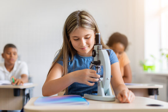 Schoolgirl using microscope during a science lesson in a bright classroom, exploring specimen while diverse classmates study at their desks in the background