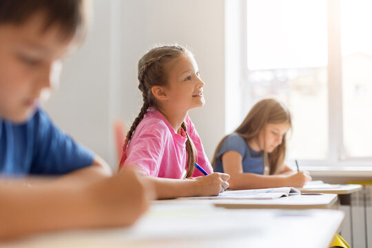 Smiling schoolgirl sitting at her classroom desk with pen and open notebook, looking up during a lesson while classmates continue writing and studying in the background