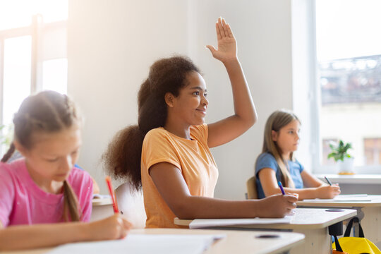 Schoolgirl raising her hand to answer a question during a classroom lesson, showing active participation while diverse classmates continue writing at their desks.