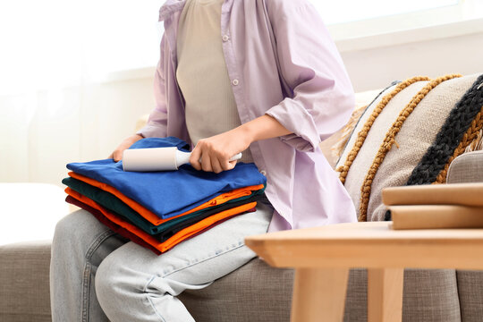 Home maintenance and clothing care, woman using a lint roller to clean a stack of folded colorful shirts while sitting on a sofa in a bright living room