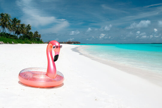 Inflatable pink flamingo pool float with sunglasses on tropical white sand beach against turquoise ocean