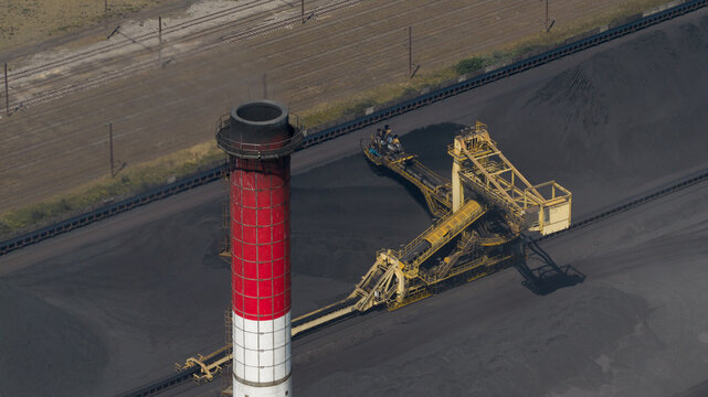 Aerial view of the ArcelorMittal industrial site featuring a tall red and white chimney, large coal storage piles, and conveyor belt systems in Dunkerque, Hauts-de-France, France.