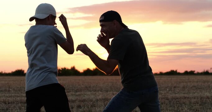 Father and son playfully practice boxing in harvested field. They spar and laugh together at sunset. Warm rural scene conveys family bonding, fitness and teamwork. Candid outdoor lifestyle footage.